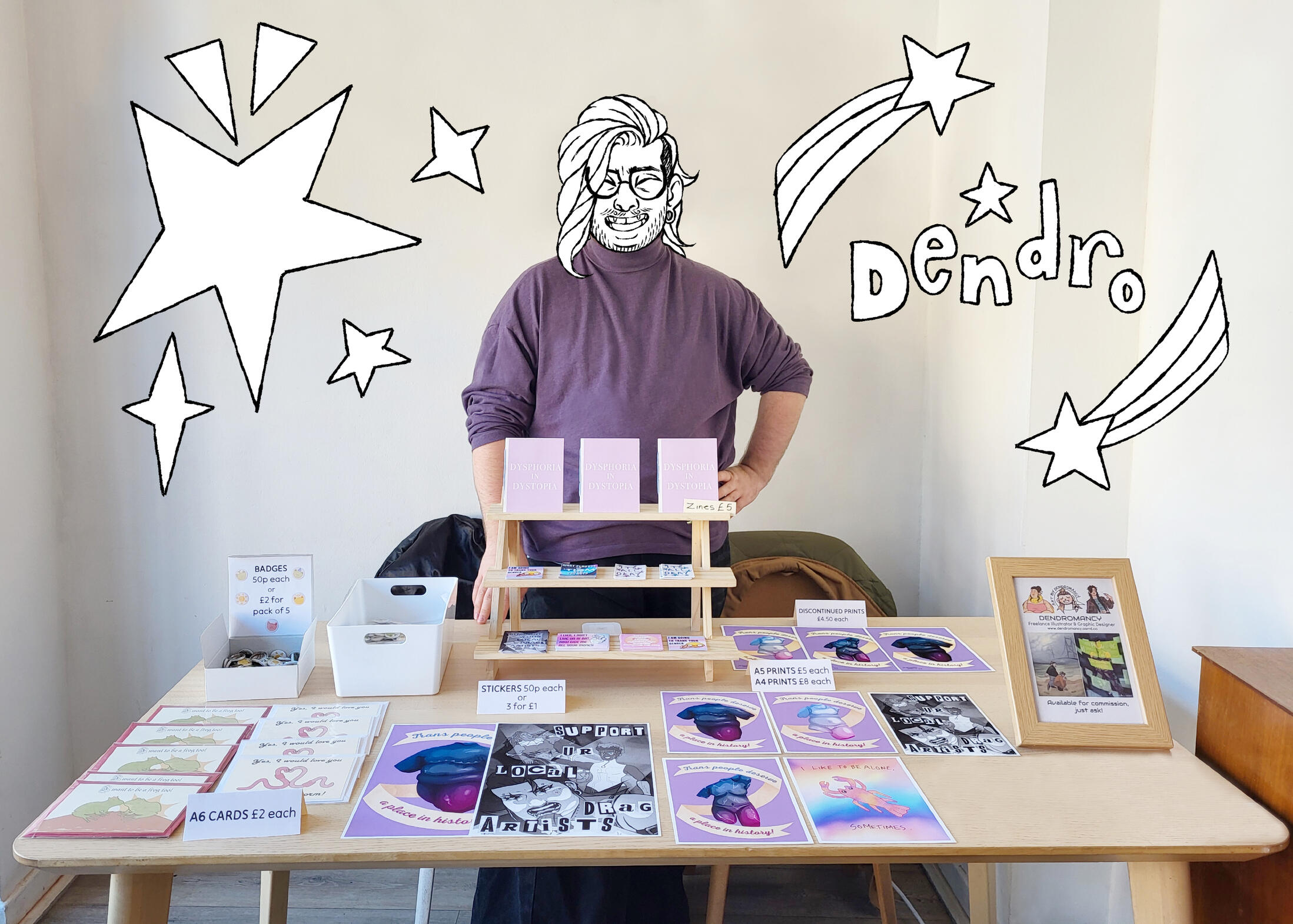 Photograph of the artist stood behind a market stall with their art on display. Their face is drawn over with an illustration of themself. There are doodles of stars around them and the word "dendro" in bubble writing.